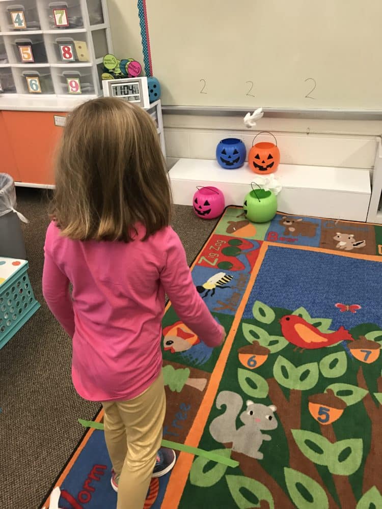 Students are tossing ghosts made from ping pong balls into plastic jack-o-laterns.