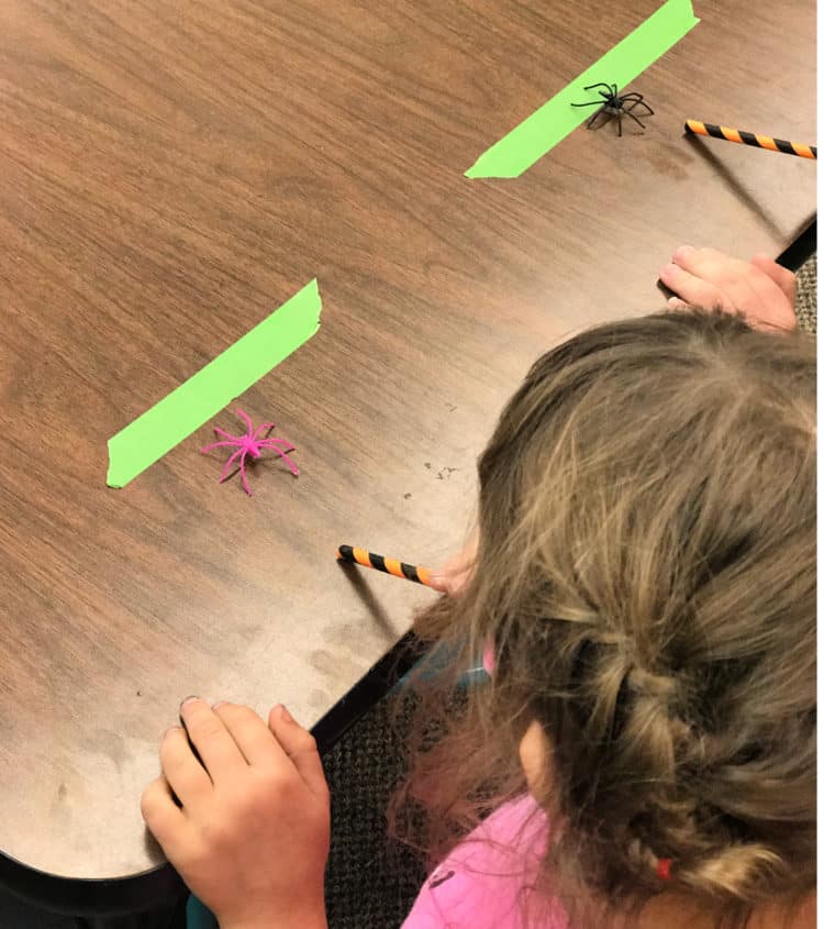 Students blow through straws and move spiders along a race track for a Halloween classroom game.