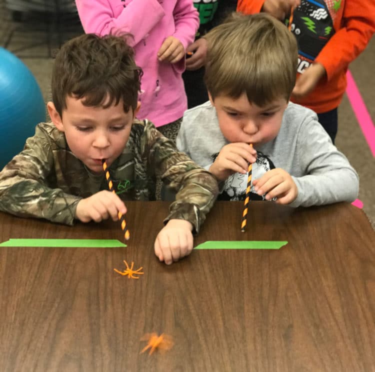 Two students are pushing plastic spiders along a race trail by blowing though straws.
